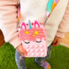 2 genți Pop It unicorn, fetiță purtând pe umăr geanta Pop It roz cu unicorn, cu curea multicoloră, pe fundal de iarbă