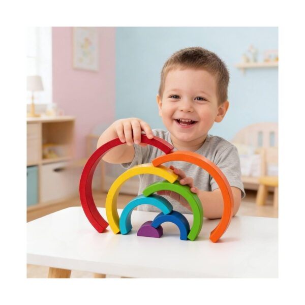 curcubeu-montessori-din-lemn-utilizare Curcubeu Montessori din lemn, băiat zâmbind la o masă albă, ridicând arcada roșie deasupra celorlalte arcade desfăcute