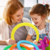Fidget Pop Tubes galben, o femeie și o fetiță întind împreună un tub fidget galben; pe masă se văd tuburi albastre, verzi