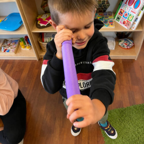 fidget-pop-tubes-xl-mov-utilizare Fidget Pop Tubes XL, băiat care ține un tub Pop Tubes mov lângă ochi, într-o cameră de joacă