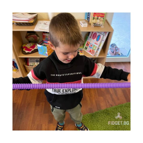 fidget-pop-tubes-xl-roz-utilizare-4 Fidget Pop Tubes XL, băiat care întinde un tub mov-roz în jurul corpului, în fața unui raft cu jucării