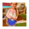 geanta-pop-it-28-26-cm-utilizare Geantă Pop It, fetiță într-o cameră de joacă, ținând în fața ei geanta Pop It curcubeu.