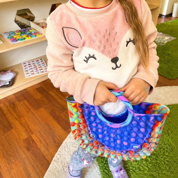 geanta-pop-it-28-26-cm-utilizare-2 Geantă Pop It, fetiță care susține geanta cu ambele mâini, cu mânerul rotund și interiorul vizibile.