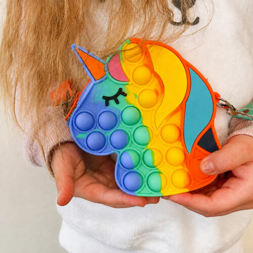 Geantă Pop It unicorn, copil care ține cu ambele mâini o geantă Pop It în formă de cap de unicorn, în culori curcubeu
