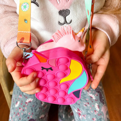 Geantă Pop It unicorn rotundă, o fetiță în bluză cu iepuraș ține cu ambele mâini o geantă Pop It fucsia în formă de unicorn