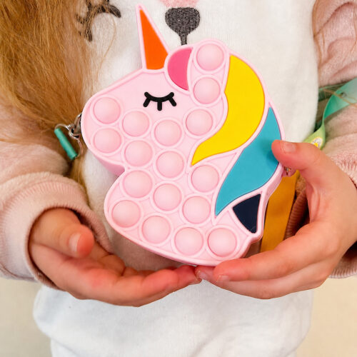 Geantă Pop It unicorn, copil în pulover alb pufos care ține geanta Pop It unicorn roz, cu ochiul închis și coama colorată