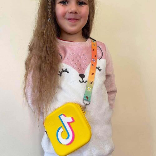 Geantă TikTok, fetiță blondă care poartă în bandulieră o geantă TikTok galbenă, cu bretea colorată imprimată
