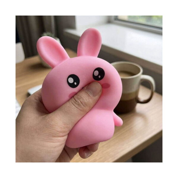 iepuras-roz-squishy-utilizare Iepuraș roz squishy, mână care strânge iepurașul squishy roz, deformându-l, pe un birou de lemn cu o cană de cafea în fundal