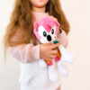 Jucărie de pluș Sonic, o fetiță cu păr lung blond-roșcat, cu fața neinclusă complet în cadru, ține în brațe jucăria roz Amy