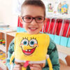 Jucărie de pluș SpongeBob, băiat cu ochelari, într-o sală de clasă cu rafturi și materiale școlare