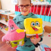 Jucărie de pluș SpongeBob, băiat cu ochelari ținând în brațe două jucării: steaua roz Patrick în stânga și SpongeBob