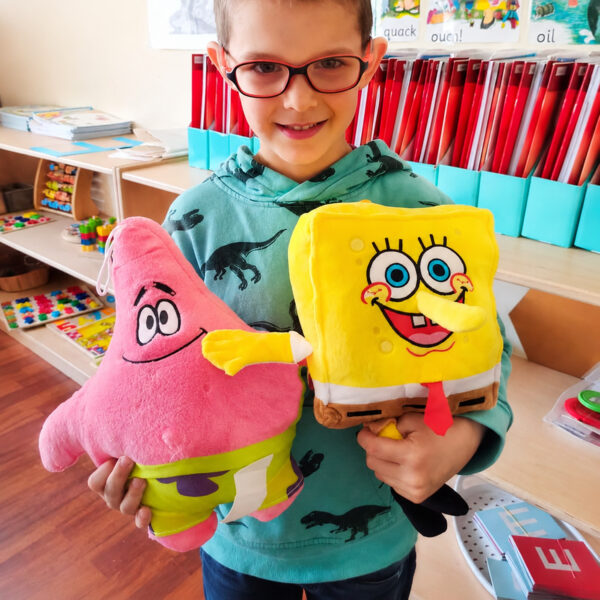 Jucărie de pluș SpongeBob, băiat cu ochelari ținând două jucării: Patrick roz în stânga și SpongeBob galben în dreapta