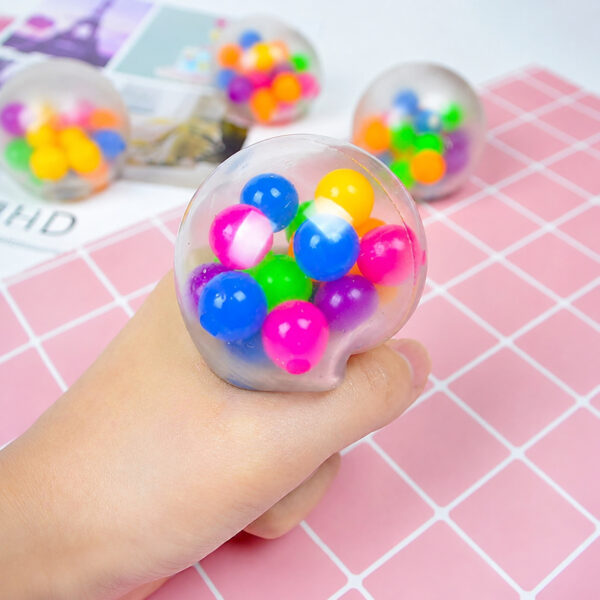 minge-squishy-3 Minge squishy, mână care strânge mingea squishy transparentă cu bile colorate, pe un fundal roz cu carouri