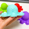 pop-it-caracatita-albastru-dimensiuni Pop It Caracatiță, mână care ține o caracatiță Pop It mentă, cu alte caracatițe verde, roșu și mov în fundal