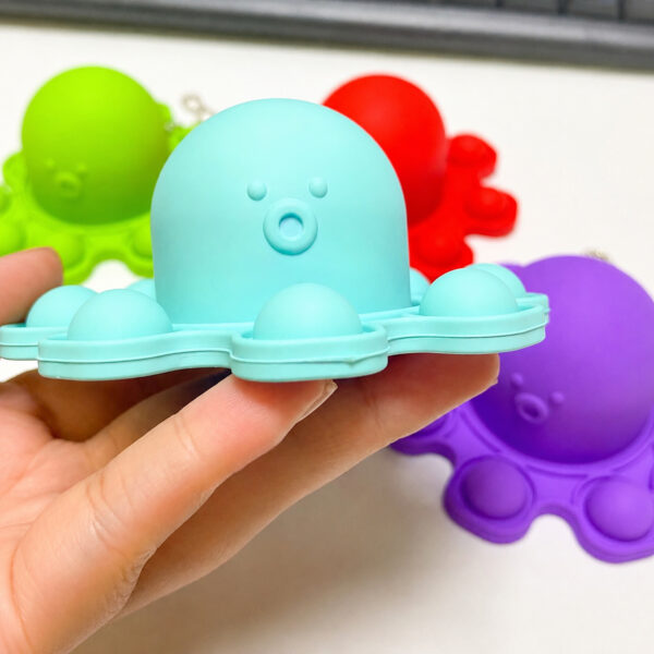 pop-it-caracatita-albastru-dimensiuni Pop It Caracatiță, mână care ține o caracatiță Pop It mentă, cu alte caracatițe verde, roșu și mov în fundal