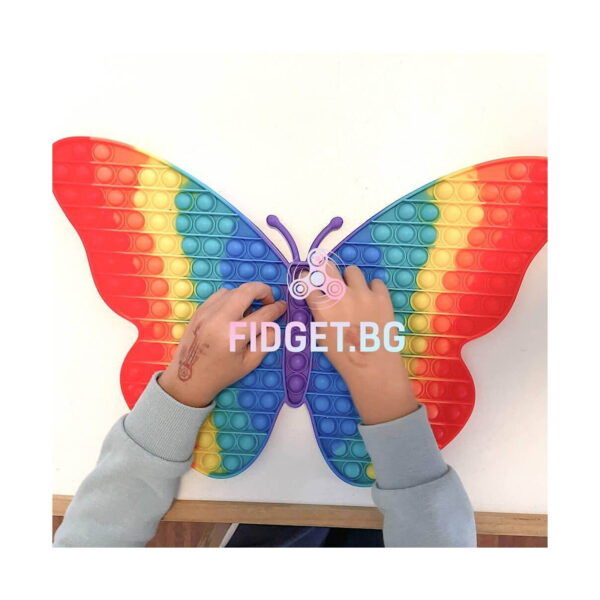 Pop It Fluture Maxi, fluturele Pop It așezat pe o masă albă, cu mâinile unui copil care apasă bulele; ALT nu menționează