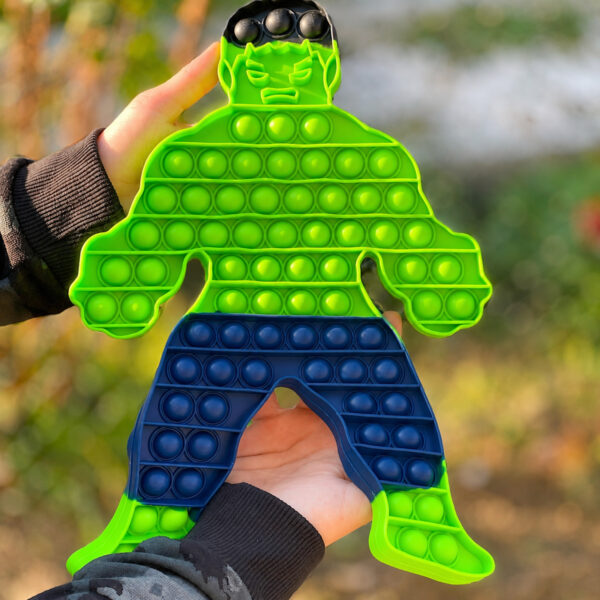 Pop It Hulk, mâinile unei persoane țin jucăria Pop It verde în formă de personaj musculos, în exterior, pe fundal cu frunziș