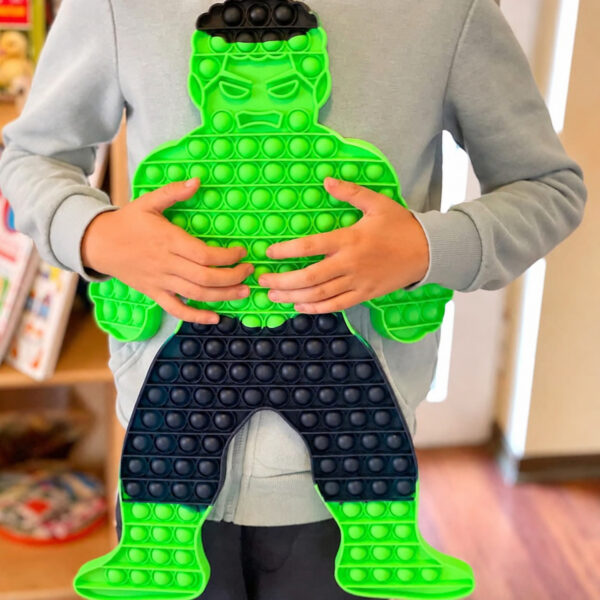 Pop It Hulk, un copil în bluză gri ține în brațe jucăria Pop It verde în formă de personaj musculos, într-o cameră