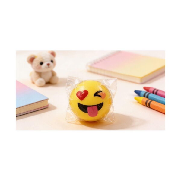 set-3-inele-emoji-luminoase-ambalaj Set 3 inele Emoji luminoase, o minge emoji galbenă ambalată în folie, așezată pe un birou lângă un ursuleț de pluș