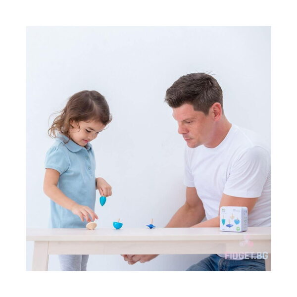 set-4-spinner-din-lemn-utilizare-3 Set 4 Spinner din lemn, fetiță și un bărbat la o masă albă, cu titireze pe masă și cutia metalică a setului în dreapta