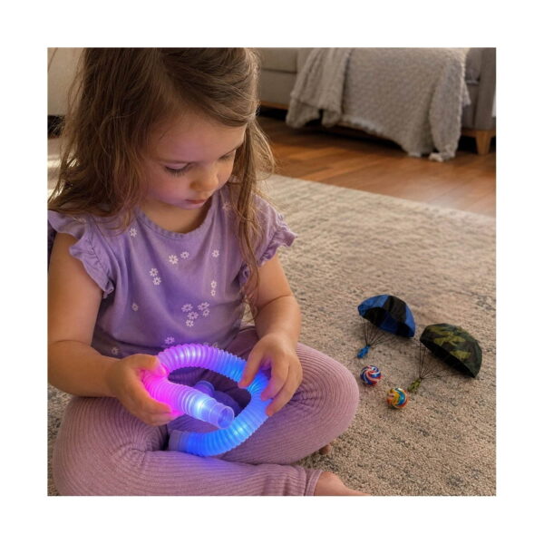 Set Aventuri Fly 11 în 1, fetiță așezată pe covor, ținând un tub luminos aprins îndoit în buclă, cu parașută și mingi în jur