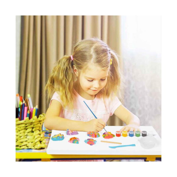 Set creativ Zâne și Unicorni, o fetiță blondă pictează la o masă figurine de zâne și unicorni; alături sunt borcănele