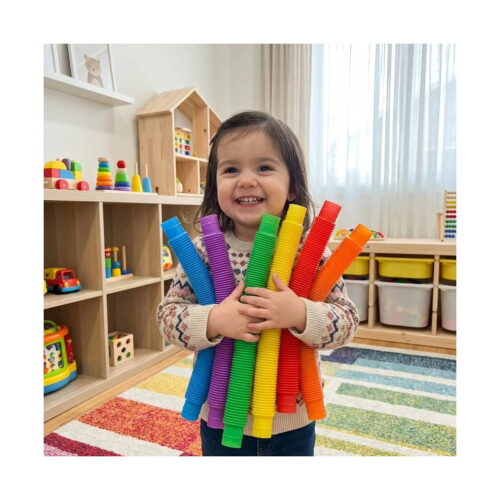 Set de 7 Pop Tubes mari, fetiță zâmbitoare într-o cameră de joacă, ținând în brațe un mănunchi de tuburi Pop Tube mari