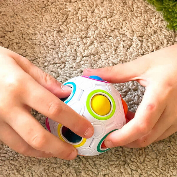 Set IQ Logică și Aventură, mâini care apasă aceeași minge albă cu butoane circulare colorate, pe un covor bej.