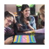 set-pop-it-utilizare-3 Set Pop It, grup de trei persoane la o masă, cu tabla Pop It pastel și două zaruri în zona centrală, cu textul POP suprapus