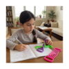 set-senzorial-iq-14-jucarii-utilizare Set senzorial IQ, fetiță care scrie într-un caiet la masă, cu trei jucării senzoriale dreptunghiulare cu fire elastice