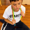 Șnur extensibil Fidget, un băiat în tricou alb, așezat pe podea, întinde cu ambele mâini mai multe șnururi Fidget colorate
