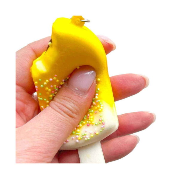 Squishy înghețată Dulce tentație, o mână strânge înghețata galbenă, materialul deformându-se sub degetul mare